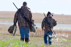 Two individuals in camouflage attire carrying shotguns walking through a grassy field near a body of water, on a duck hunting trip.