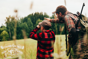 Two people in camouflage hats and plaid shirts stand in a grassy field; one looks through binoculars while the other points ahead. "Waterfowl Unlimited Guide Service" logo is at the bottom left.