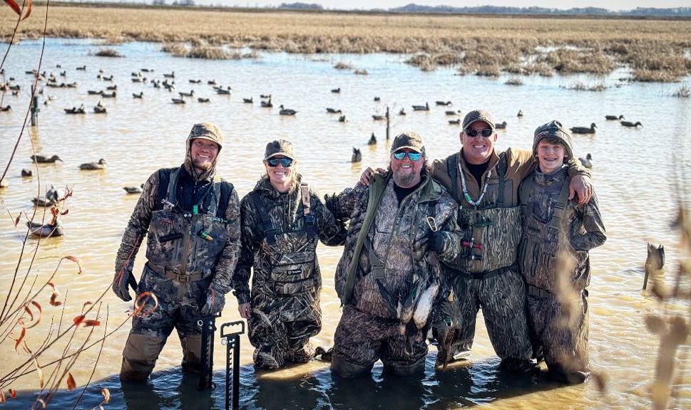 Five people in camouflage gear stand in water, smiling while holding hunting equipment. Ducks float in the background.