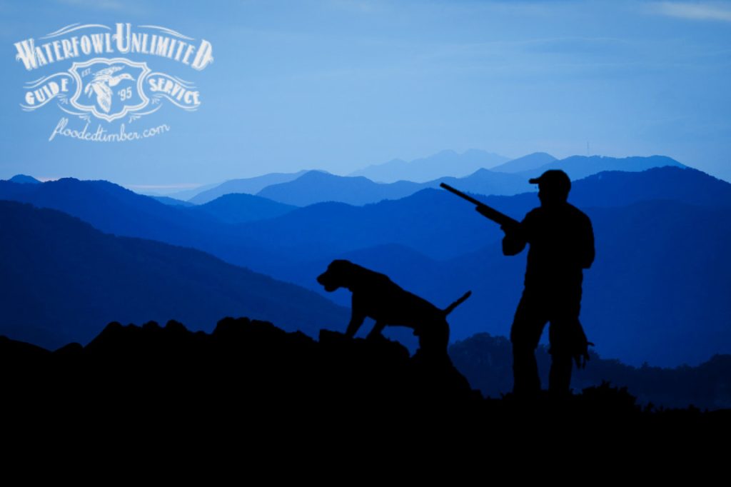 A person with a rifle stands beside a dog on a ridge, silhouetted against layered blue mountains at dusk. Waterfowl Unlimited Guide Service logo is in the top left corner.