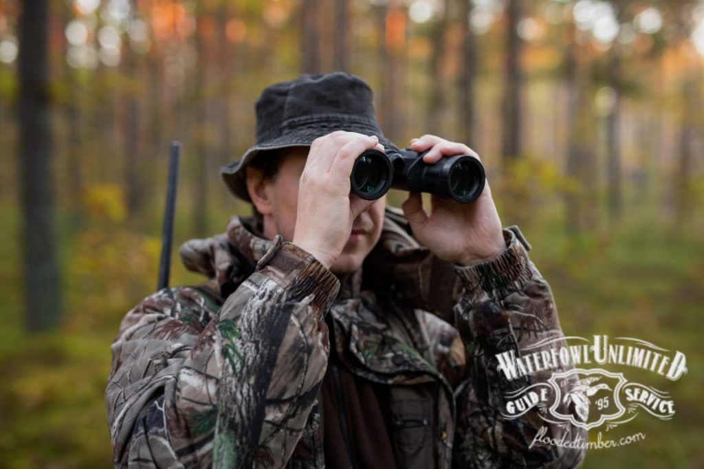 A person in camouflage clothing and a bucket hat uses binoculars in a wooded area, with a rifle strap visible over their shoulder. "Waterfowl Unlimited Guide Service" logo is in the corner.