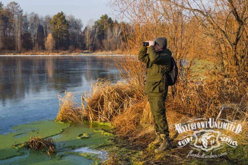 A person in outdoor gear stands on a grassy bank near a partially frozen lake, looking through binoculars with trees visible in the background.
