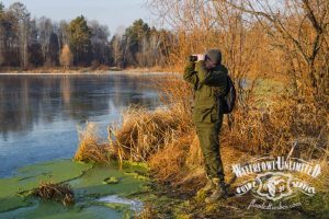 A person in outdoor gear stands on a grassy bank near a partially frozen lake, looking through binoculars with trees visible in the background.