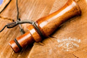 A wooden duck call with a black lanyard rests on a wooden surface next to a "Waterfowl Unlimited Guide Service" logo.