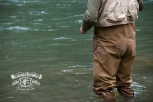 Person in chest waders stands in a river while fly fishing, holding a fishing line. Waterfowl Unlimited Guide Services logo is visible on the image.
