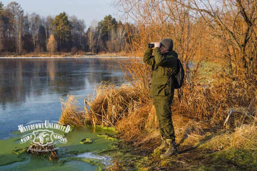 A person in outdoor gear stands by a partially frozen lake, looking through binoculars, surrounded by dry grasses and trees.