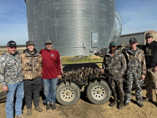 Six men in outdoor clothing stand in front of a metal grain bin and a trailer displaying a row of hunted birds.