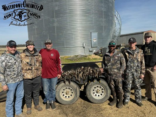 Six men in camouflage and outdoor clothing stand by a trailer displaying a row of hunted ducks, with a large metal grain bin in the background.