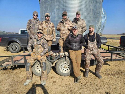 Seven men in outdoor hunting gear pose in front of a metal structure and two trucks; several ducks are displayed on the trailer in front of them.