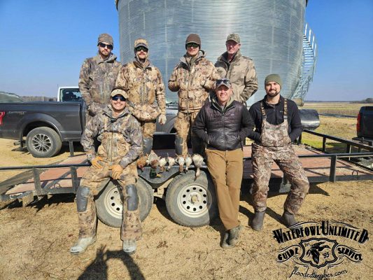 Seven men in hunting gear pose in front of a large metal tank and two trucks, with several ducks displayed on a trailer and a "Waterfowl Unlimited" logo in the corner.