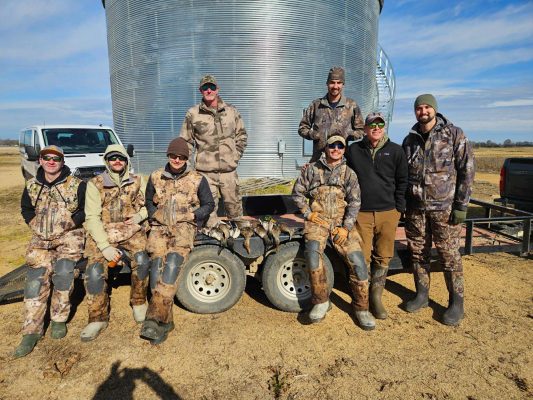 Eight people in camouflage and outdoor gear pose for a group photo in front of a large metal grain bin and a parked van on a clear day.