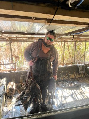 A man wearing waders and sunglasses stands in a boat holding several ducks; a black dog is by his side. Tall grasses are visible through the boat’s open sides.