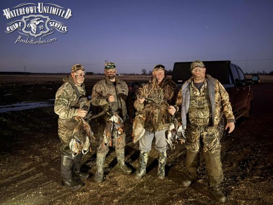 Four hunters in camouflage stand in front of a truck at dusk, each holding several ducks. A "Waterfowl Unlimited Guide Service" logo is in the top left corner.