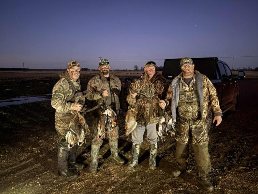 Four people in camouflage hunting gear stand outdoors at dusk holding ducks and other game birds in front of a parked truck.