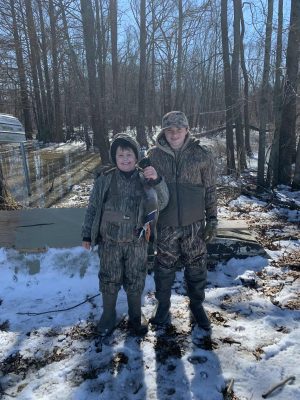Two boys in camouflage clothing stand on snowy ground in a wooded area, one holding a duck. Flooded trees and a partially submerged boat are visible in the background.