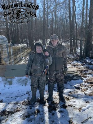 Two children dressed in camouflage hunting gear stand in a snowy, wooded area; one is holding a duck. The Waterfowl Unlimited logo appears at the top of the image.