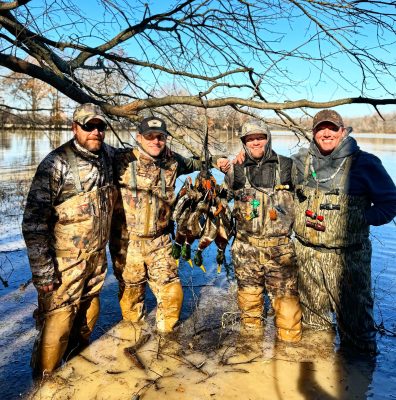 Four men in camouflage waders stand in a flooded area holding several ducks, with bare tree branches and water in the background.