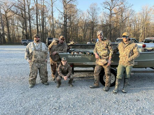 Five people in camouflage clothing pose by a small boat labeled “SeaArk” on a trailer in a gravel parking area, with leafless trees and several vehicles in the background.