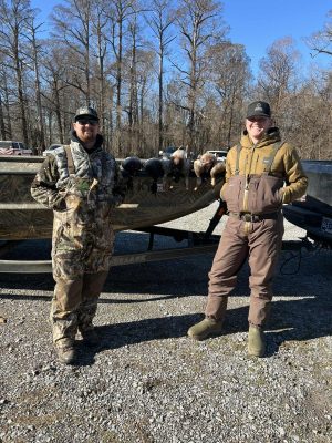 Two people in outdoor gear stand beside a camouflaged boat with several ducks laid out on top; leafless trees and vehicles are visible in the background.