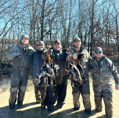 Five people in camouflage hunting gear pose outdoors, holding several ducks. They stand together in a muddy area with leafless trees in the background.