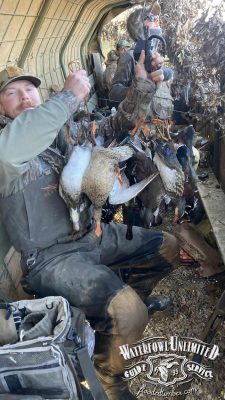 A group of hunters in camouflage sit inside a duck blind, holding several harvested ducks. "Waterfowl Unlimited Guide Service" logo is visible in the lower right corner.