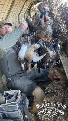 A group of hunters in camouflage sit inside a duck blind, holding several harvested ducks. "Waterfowl Unlimited Guide Service" logo is visible in the lower right corner.