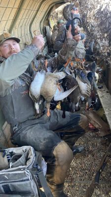 Two people in camouflage hunting gear sit in a blind, holding up several ducks they have hunted, with equipment and bags visible around them.