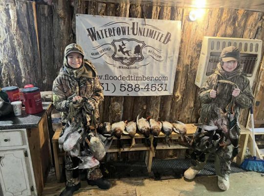 Two people in camouflage clothing stand indoors holding harvested ducks, with more ducks laid out on a bench in front of a "Waterfowl Unlimited" banner.
