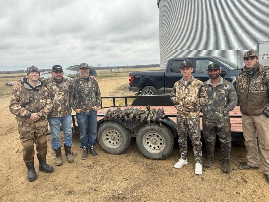 Six men in camouflage hunting gear stand beside a trailer holding several ducks, with pickup trucks and a large metal silo in the background.