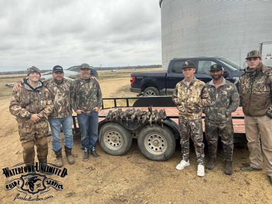 Six men in camouflage clothing stand beside a trailer with harvested ducks, a black pickup truck, and a large metal grain bin in the background.