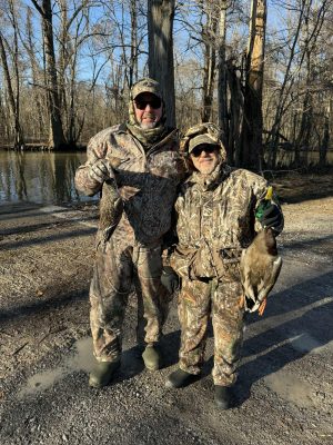 Two people in camouflage hunting gear stand on a dirt path by water, each holding a duck, surrounded by leafless trees.