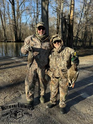 Two people in camouflage clothing stand outdoors in a wooded area, each holding a duck. The "Waterfowl Unlimited" logo is visible in the lower left corner.