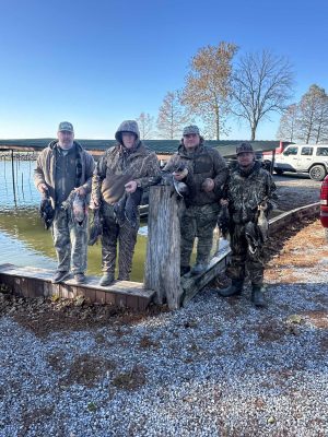 Four people in camouflage clothing stand by a lakeside holding several ducks, with a truck and bare trees visible in the background.