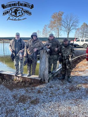 Four hunters in camouflage stand by a waterfront holding ducks, with trucks and trees in the background. A "Waterfowl Unlimited Guide Service" logo is at the top of the image.