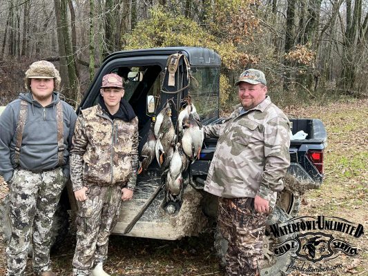 Three people in camouflage stand by an off-road vehicle displaying several hunted ducks hanging from the side; a wooded area is in the background.