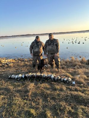 Two hunters in camouflage stand beside a black dog and a row of hunted ducks on the ground near a wetland with decoys in the water.