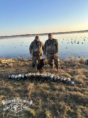 Two hunters in camouflage stand by a black dog near a row of harvested ducks on grass beside a lake, with decoys in the water and a logo in the bottom left corner.