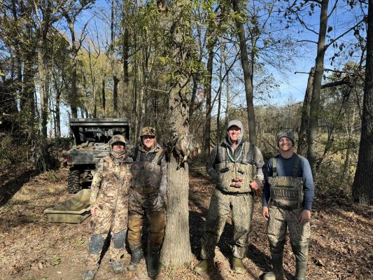 Four people in camouflage hunting gear stand outdoors by a tree with ducks hanging from it; an ATV is parked nearby in a wooded area.