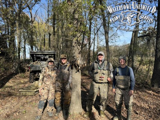Four people in camouflage and waders stand in a wooded area next to a tree with several ducks hanging from it; an ATV and the “Waterfowl Unlimited Guide Service” logo are visible.
