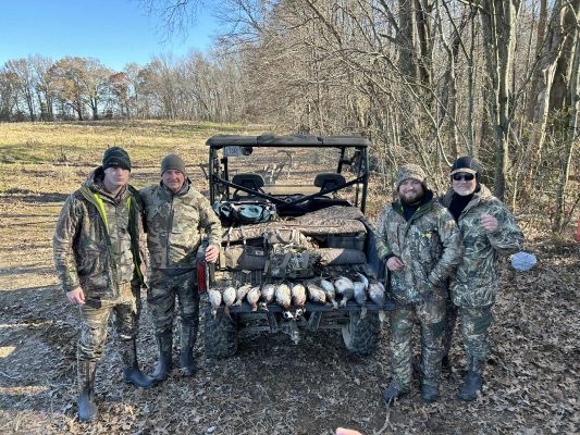 Four people in camouflage hunting gear stand beside an off-road vehicle with several ducks laid out on its back, in a wooded outdoor setting.