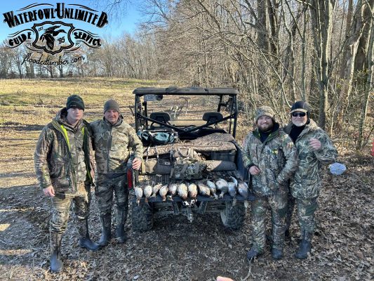 Four people in camouflage stand by an off-road vehicle displaying a row of hunted ducks. The scene is outdoors in a wooded area with fallen leaves on the ground.