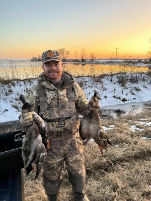 A man in camouflage hunting gear stands outdoors at sunset, holding two ducks, with snow-covered ground and a stream in the background.