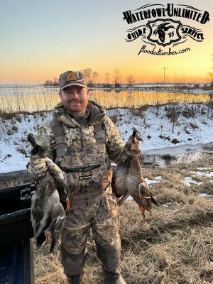 A man in camouflage hunting gear holds two ducks outdoors near a snowy riverbank at sunset, with a Waterfowl Unlimited Guide Service logo above.