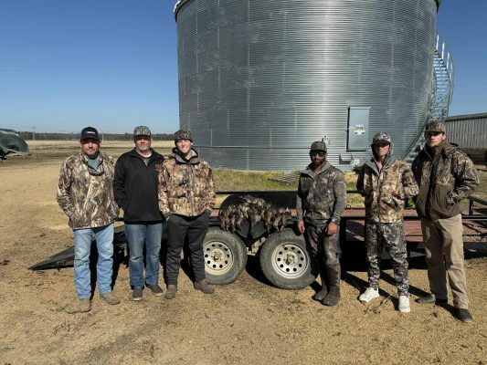 Six men in camouflage clothing stand in front of a metal grain silo and trailer outdoors on a sunny day.