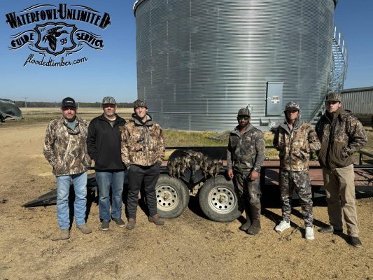 Six men in camouflage and outdoor clothing stand in front of a trailer with harvested game birds, beside a large metal grain bin on a dirt lot.