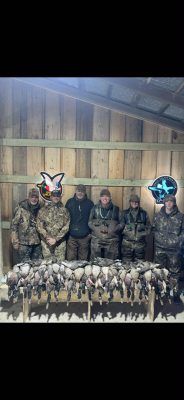 Five hunters in camouflage pose indoors behind a display of numerous harvested ducks hanging on a wooden rack, with wooden walls and hunting-themed neon signs in the background.