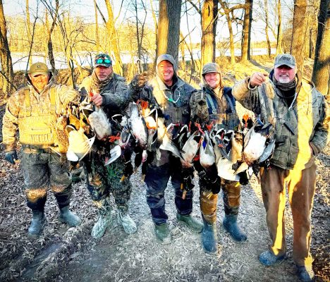 Five people in outdoor gear stand in a wooded area holding multiple ducks, likely after a successful hunting trip, with bare trees and sunlight in the background.