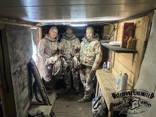 Three people in camouflage hunting gear stand in a wooden cabin holding ducks. Hunting supplies and condiments are visible on shelves. "Waterfowl Unlimited" logo on lower right corner.