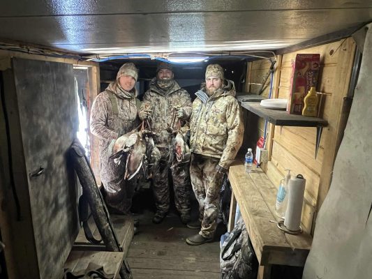 Three people in camouflage clothing stand in a wooden hunting cabin holding ducks. Shelves with condiments, paper towels, and a water bottle are visible on the right.