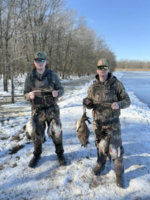 Two people in camouflage gear stand on snow by a frozen pond, each holding several ducks, with bare trees in the background.